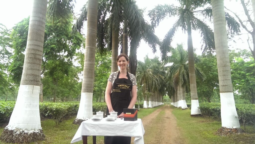 IMAG2900 woman in Twinings apron standing behind a table with tea brewing equipment, in a tea garden