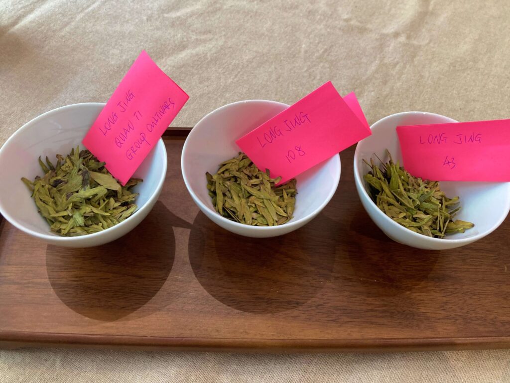 Three white bowls of loose leaf Longjing Tea, each labeled with pink notes reading Long Jing Qiang Ti Extra Quality, Long Jing 108, and Long Jing 43—iconic tea cultivars—sit on a wooden tray atop a beige surface.