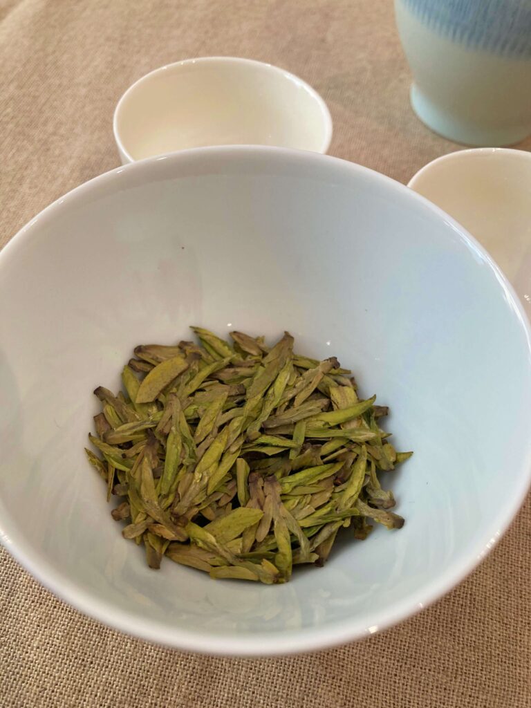 A white bowl filled with loose, flat Longjing Tea leaves sits on a beige tablecloth next to two empty white teacups and a partially visible teapot—an inviting scene featuring this iconic tea among celebrated tea cultivars.