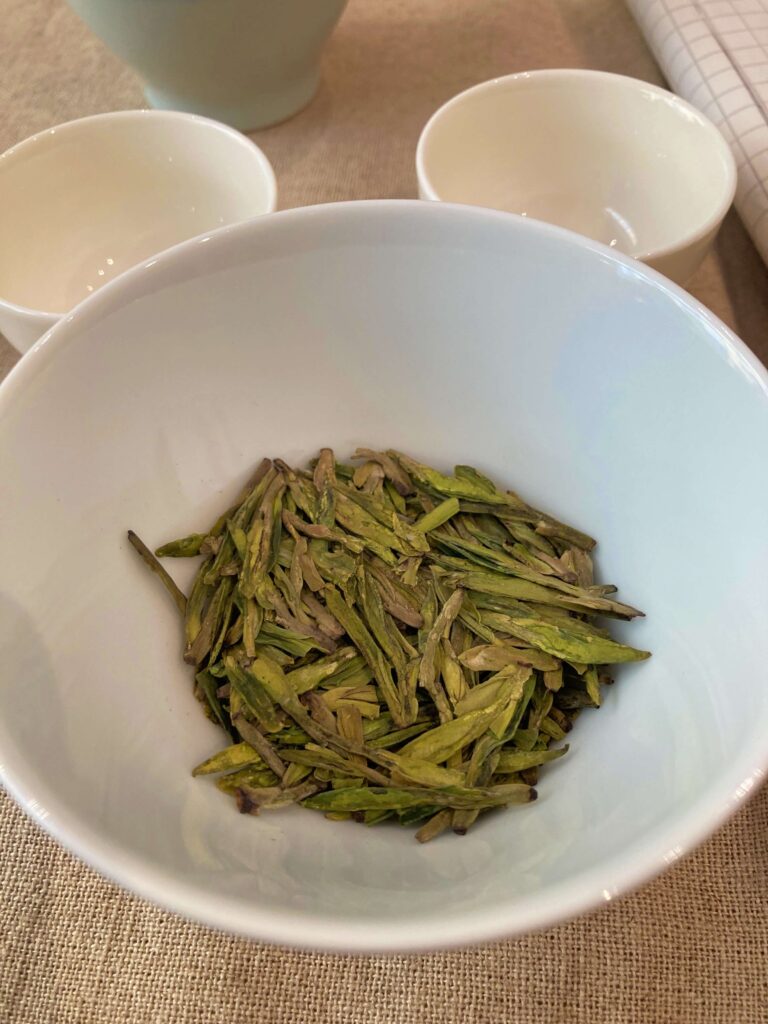 A white bowl filled with loose, flat Longjing tea leaves—an iconic Chinese tea—sits on a beige surface, with two empty white cups and part of a teapot visible in the background.