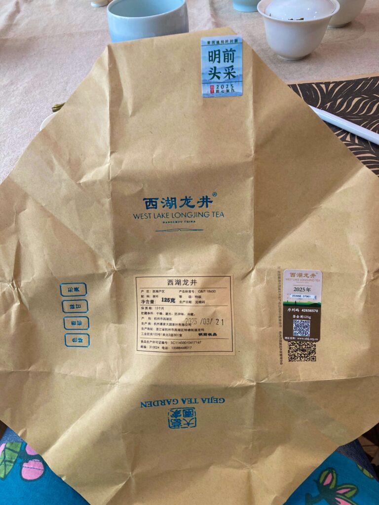 A brown West Lake Longjing tea wrapper, highlighting this iconic tea with Chinese and English text, QR codes, and labels, is spread out on a table near white teacups and a floral-patterned cloth.
