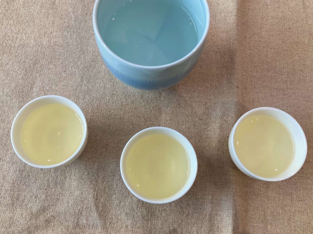 Three small cups of pale yellow Longjing tea are arranged in a row below a larger bowl of water, all set on a beige fabric surface.