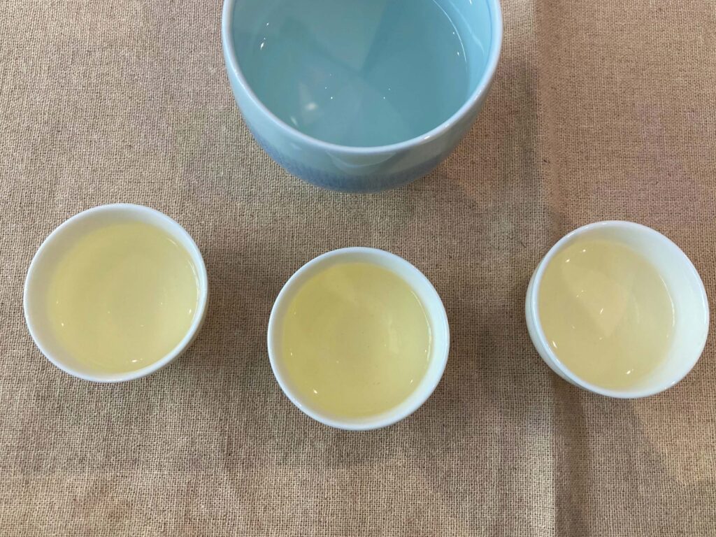 Three small cups filled with light yellow Longjing Tea, an iconic Chinese tea, are arranged in a row below a larger blue bowl of water, all placed on a tan fabric surface.