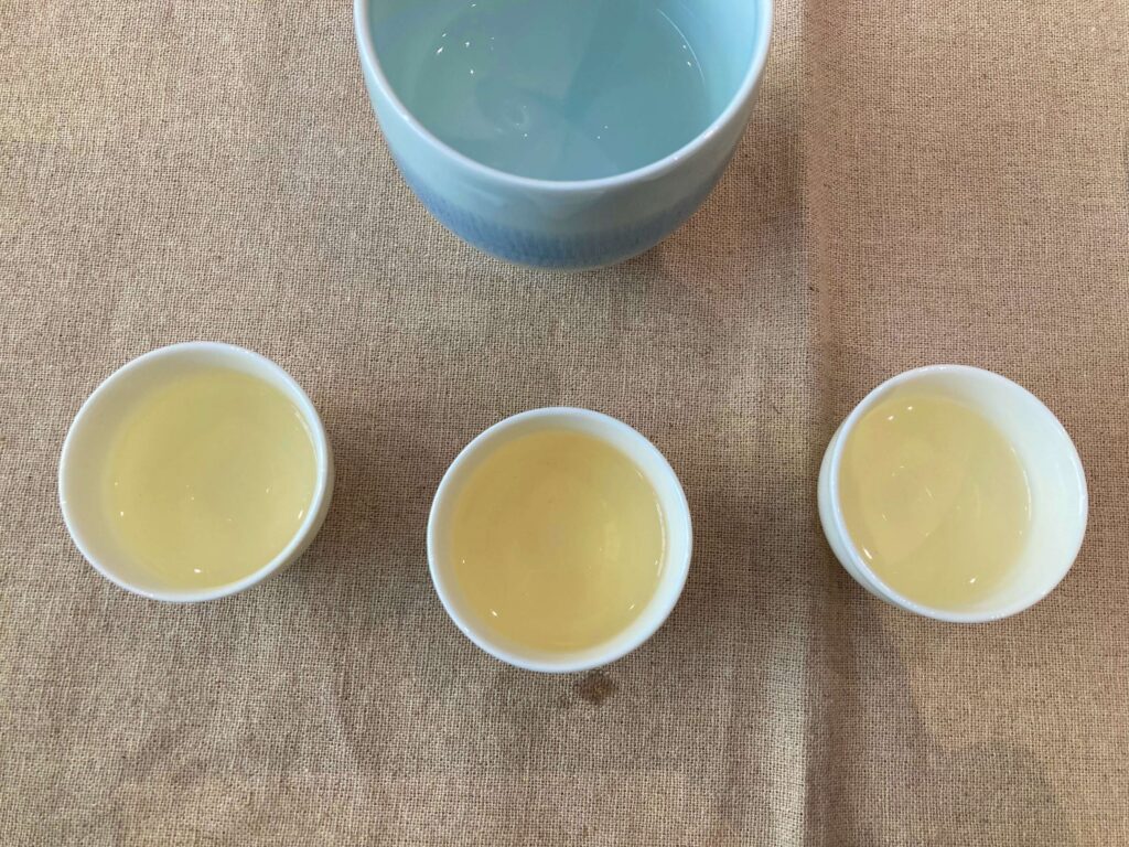Three small cups filled with pale yellow Longjing tea are arranged in a row on a beige cloth, suggesting a tea comparison, with a larger blue bowl partially visible above them.