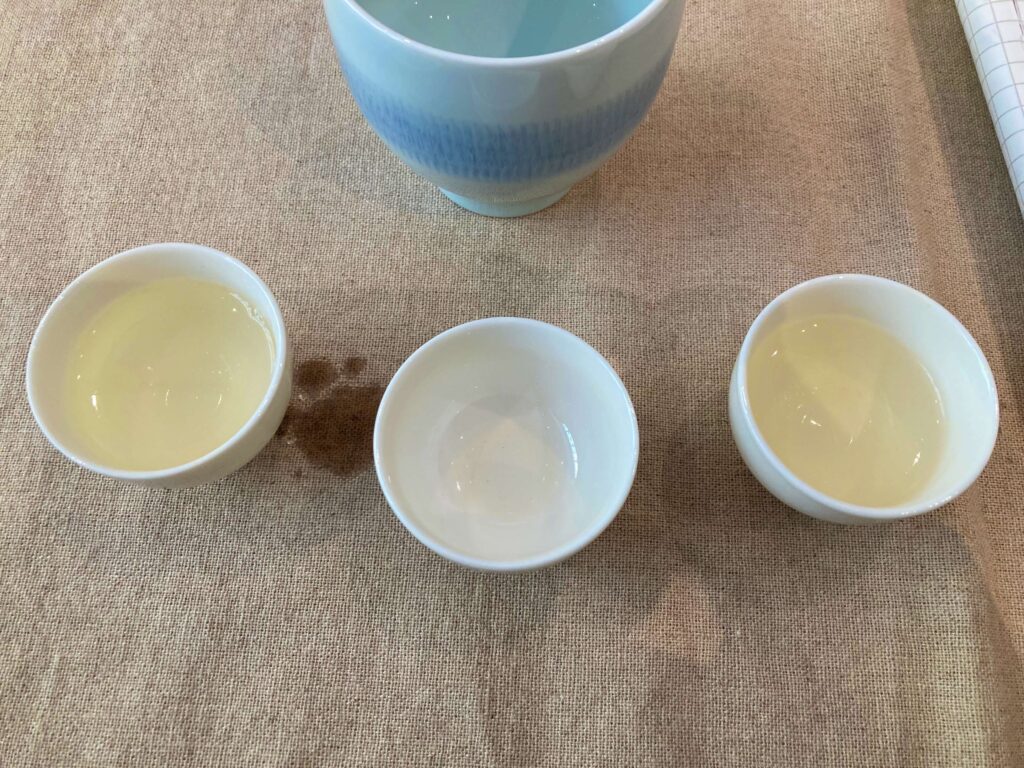 Three small white cups are arranged in a row on a beige cloth. The left and right cups hold light yellow Longjing Tea, while the center cup is empty. A larger blue-and-white cup, hinting at this iconic tea’s rich tradition, is partially visible above them.