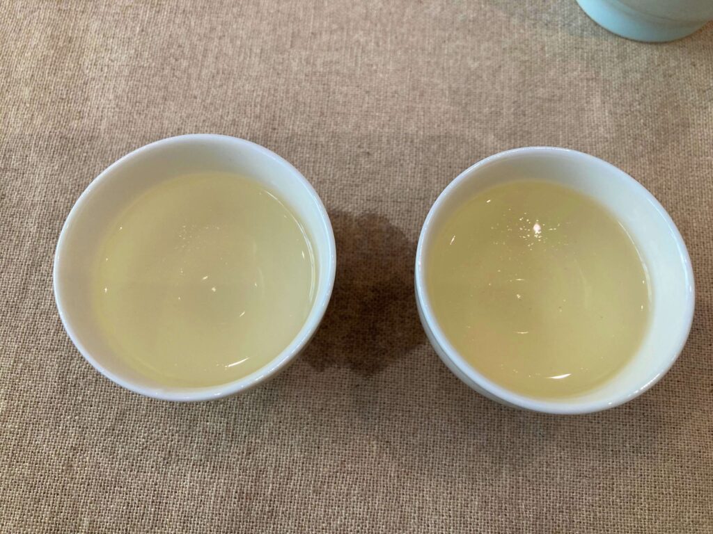 Two white ceramic cups filled with pale yellow Longjing Tea are placed side by side on a beige fabric surface, with a small water stain visible between them.