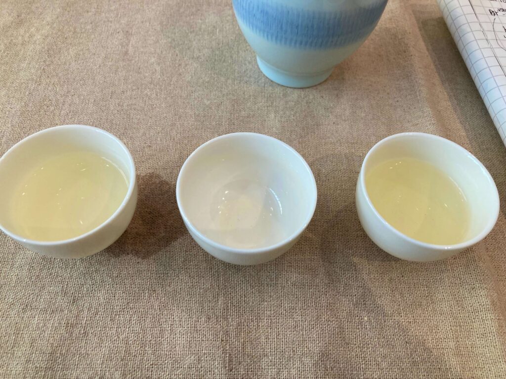 Three small white cups, two filled with iconic Chinese Longjing tea and one empty, are arranged in a row on a beige cloth. A partially visible bowl and a notebook are in the background.
