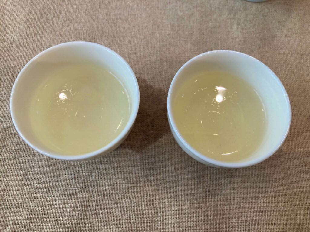 Two white cups filled with light yellow Longjing tea are placed side by side on a beige fabric surface. Bright reflections are visible on the surface of the tea in each cup, suggesting a subtle tea comparison.