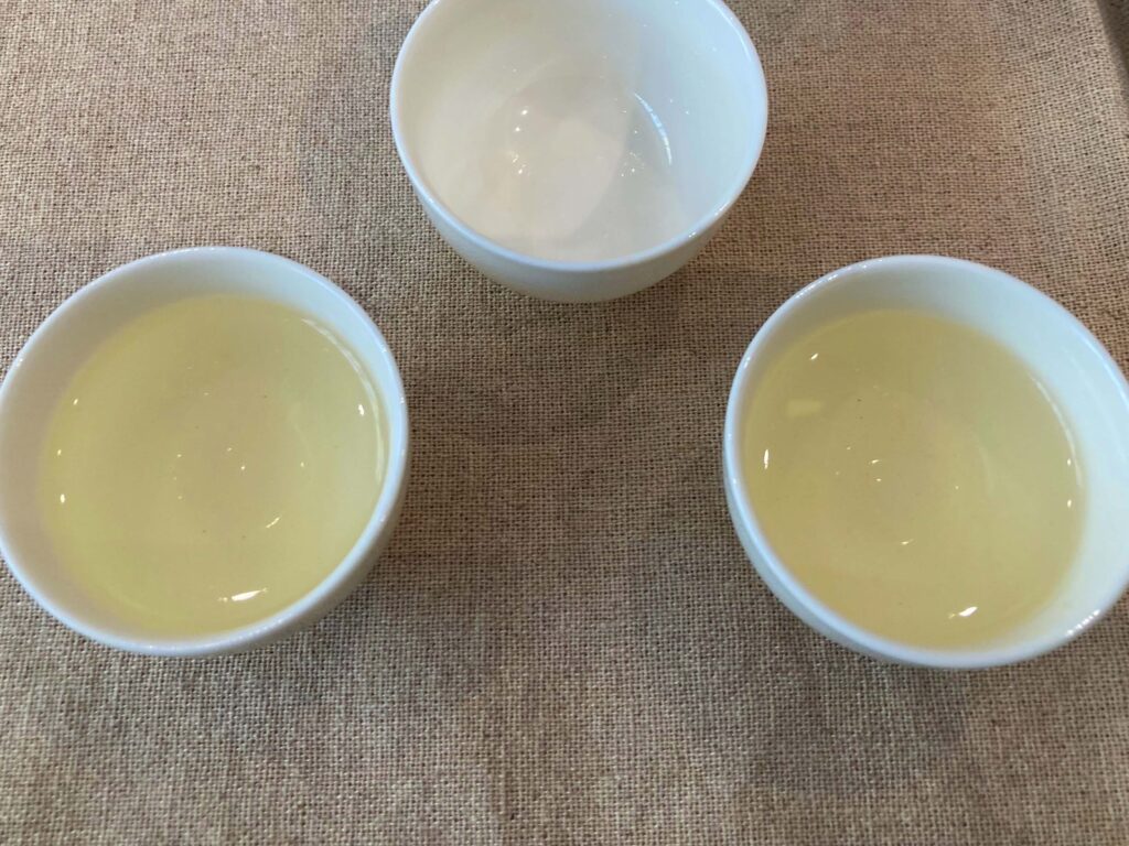 Three small white bowls sit on a beige fabric surface; two bowls hold a light yellow liquid of Chinese green tea, likely brewed from Longjing cultivars, while the third bowl in the center remains empty.