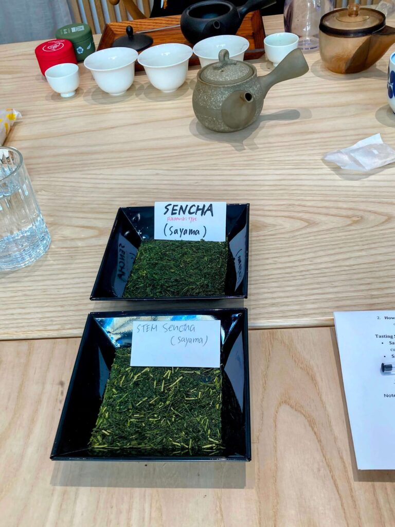 Two black trays on a wooden table showcase loose Sayama Sencha and Stem Sencha—premium Japanese tea varieties. A teapot, cups, glass of water, and tea utensils complete this elegant setup.
