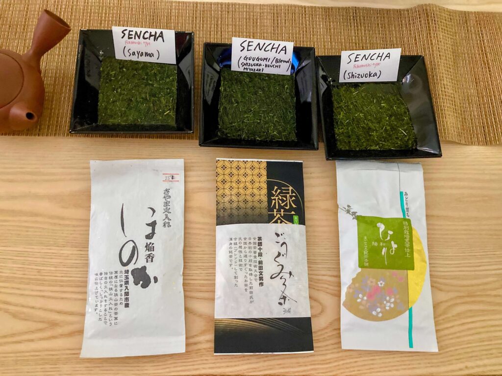 Three square dishes with different varieties of loose green tea labeled “Sencha” are displayed on a wooden surface, with three corresponding tea packages and a brown teapot partially visible on the left side.