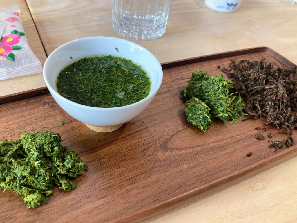 A white bowl of Sayama Sencha green tea sits on a wooden tray, surrounded by piles of loose green and brown tea leaves. A glass of water and a floral-patterned napkin complete this Japanese Tea Tasting scene.
