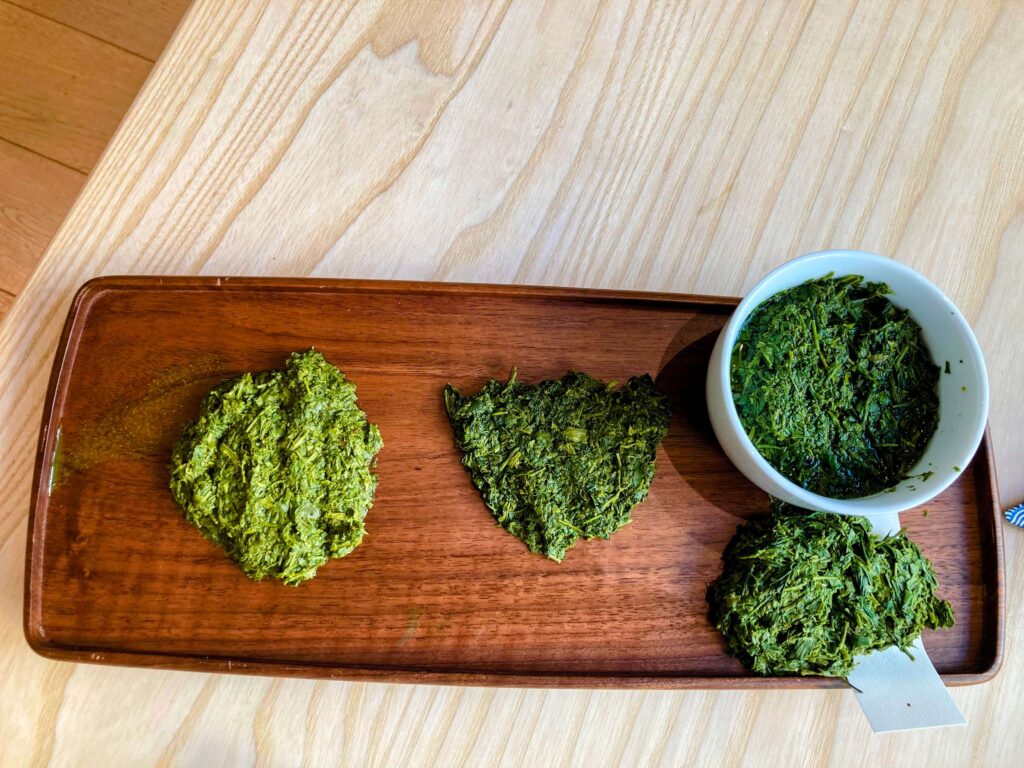 A wooden tray holds three piles of wet sencha tea leaves, with a small white bowl filled with the same tea on the side. The tray rests on a light wooden table.