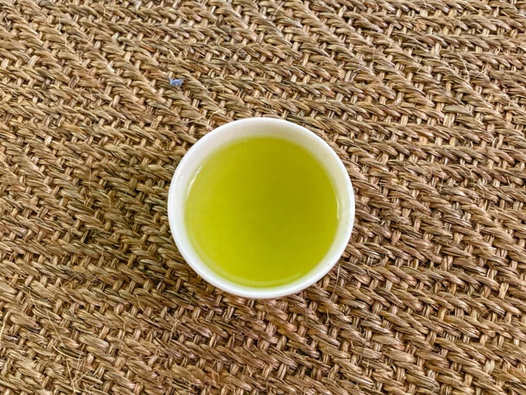 A white cup filled with green tea sits on a woven, textured brown mat, viewed from above.