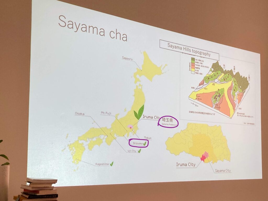 A presentation slide about Sayama cha features maps of Japan highlighting Iruma City and the Sayama Hills topography, with labeled areas and a small stack of books visible in the bottom left corner.