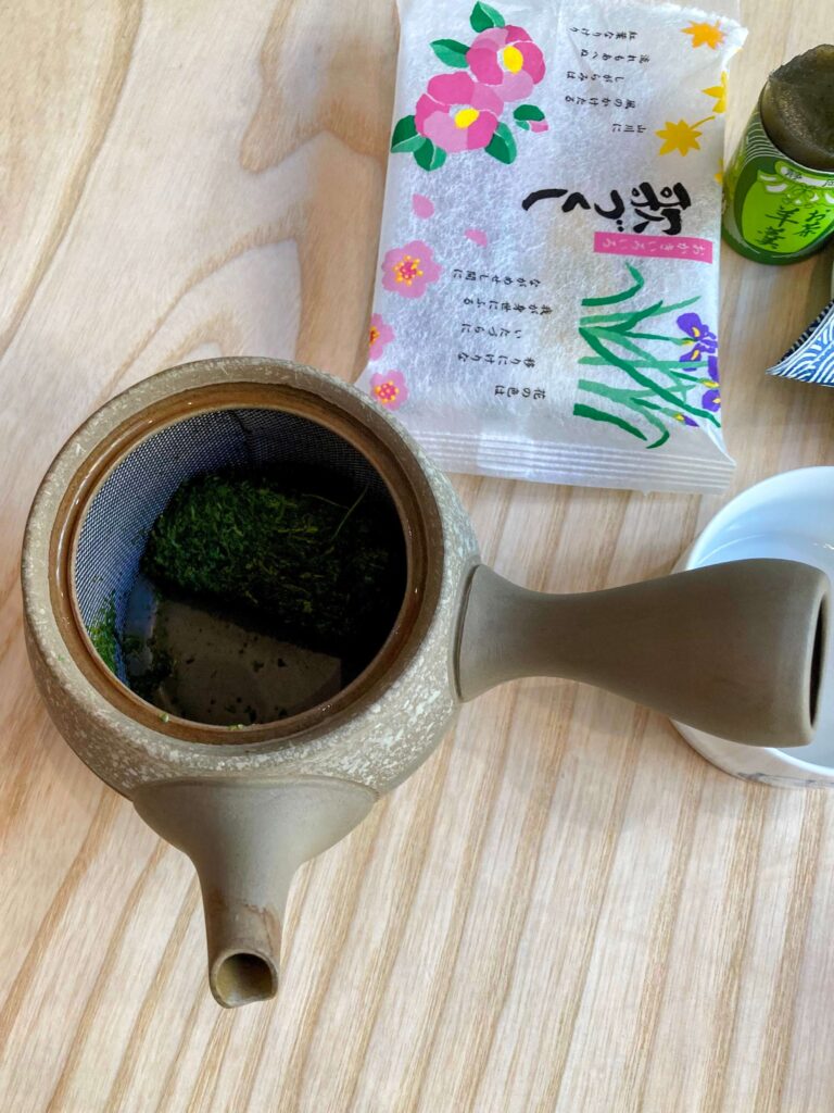 A Japanese teapot with green tea leaves inside sits on a light wooden table beside a white teacup, a colorful tea package with floral designs, and a small container of matcha.