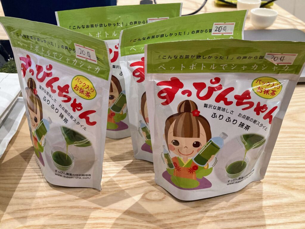 Three green and white packets of Japanese tea with a cartoon girl in a kimono on the front, placed on a wooden table. Each packet has a price tag of 204 yen.