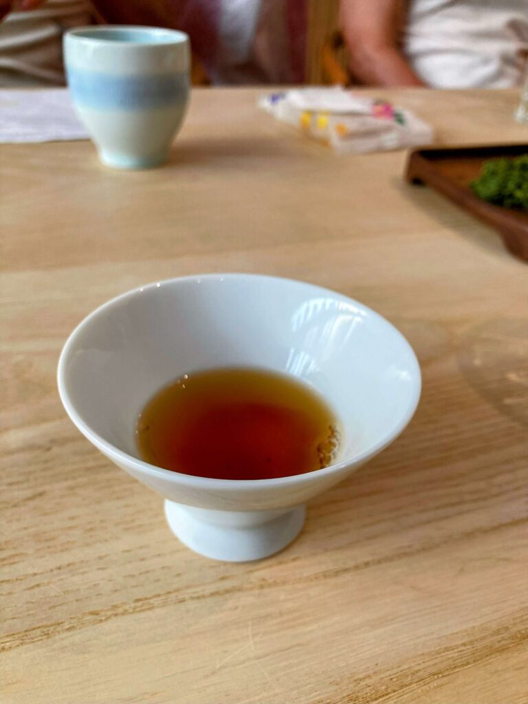 A small white teacup with brown Japanese black tea sits on a light wooden table; in the background are another cup, some packets, and people enjoying a Japanese tea tasting.