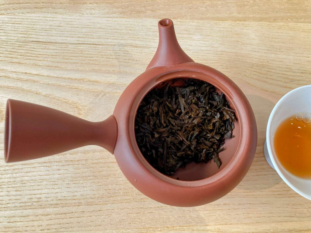 A close-up of a clay teapot filled with loose black tea leaves, placed on a light wooden surface next to a white cup containing amber-colored tea—a perfect scene for Japanese Tea Tasting.