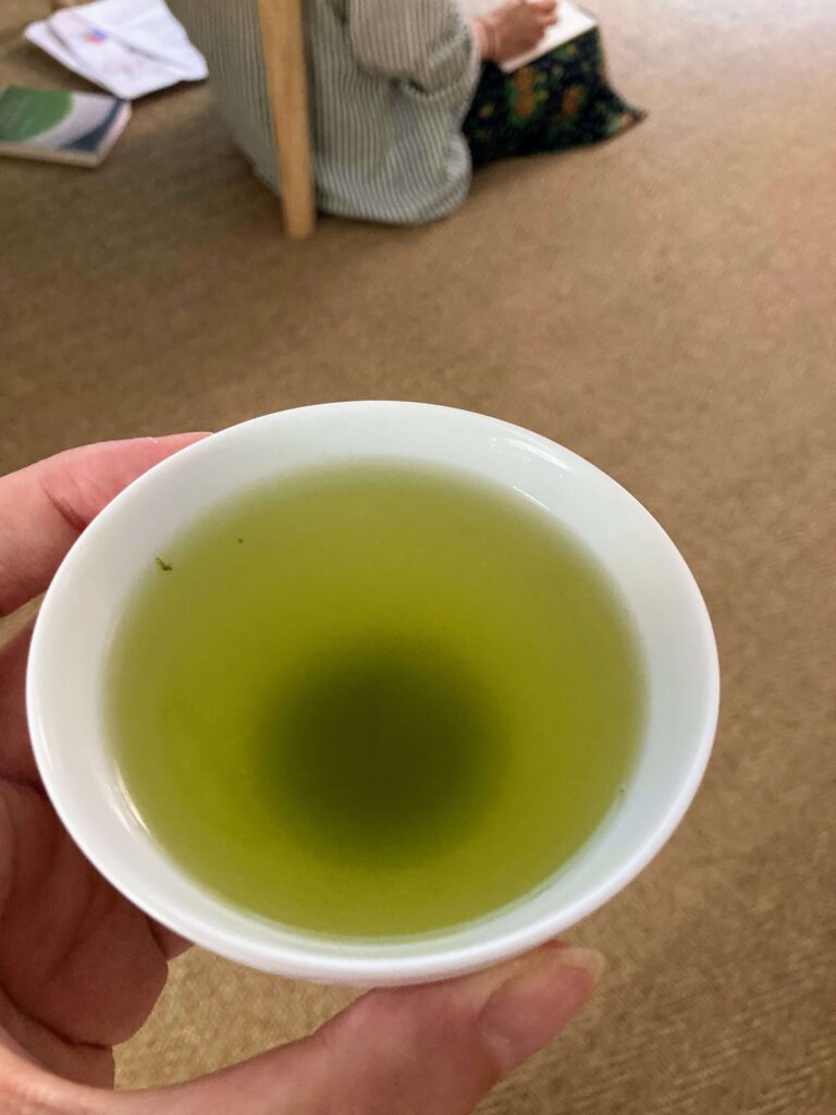 A hand holds a white cup filled with vibrant Sayama Sencha. In the background, a person sits on the floor, partially visible, next to some books or papers. The setting exudes the calm of a Japanese tea tasting.