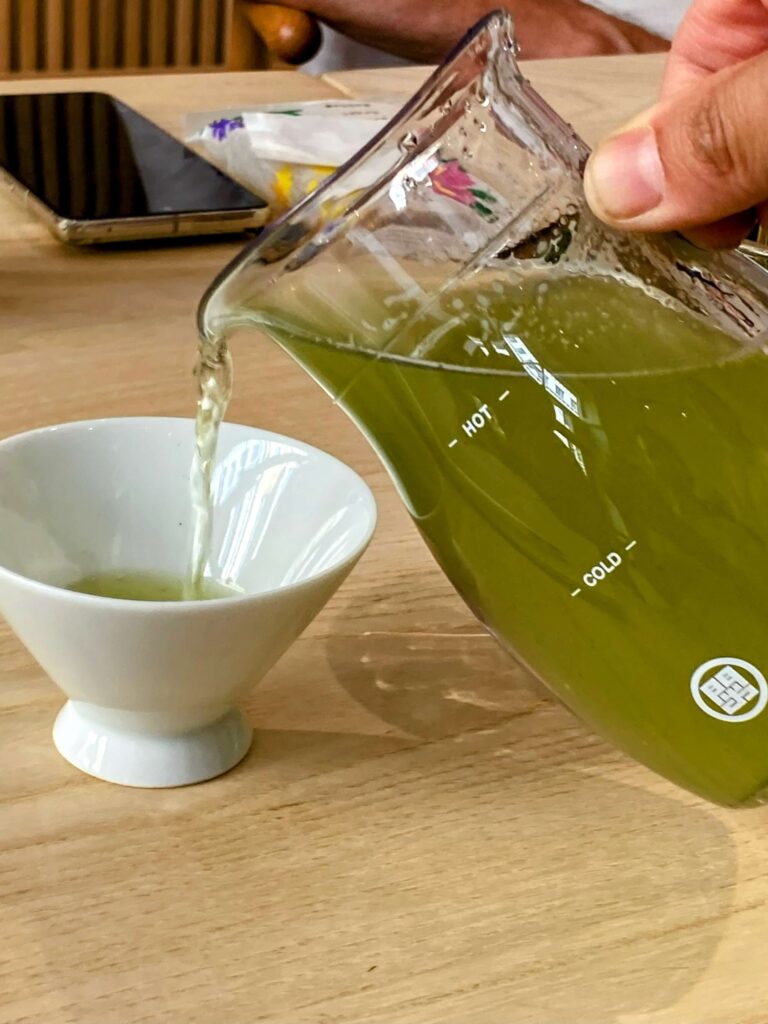 A hand pours cold brew Sayama Sencha green tea from a glass pitcher into a white teacup on a wooden table; other items are in the background, evoking an authentic Japanese Tea Tasting scene.