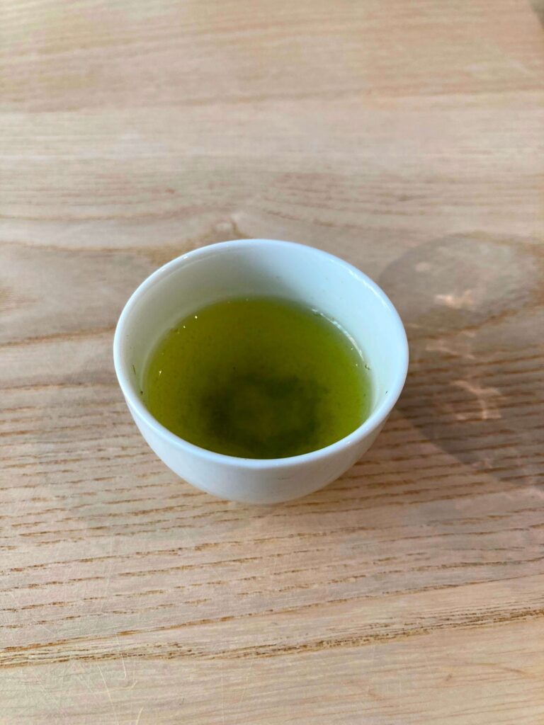 A small white cup filled with Sayama Sencha green tea sits on a light wooden surface, perfect for a Japanese Tea Tasting experience.