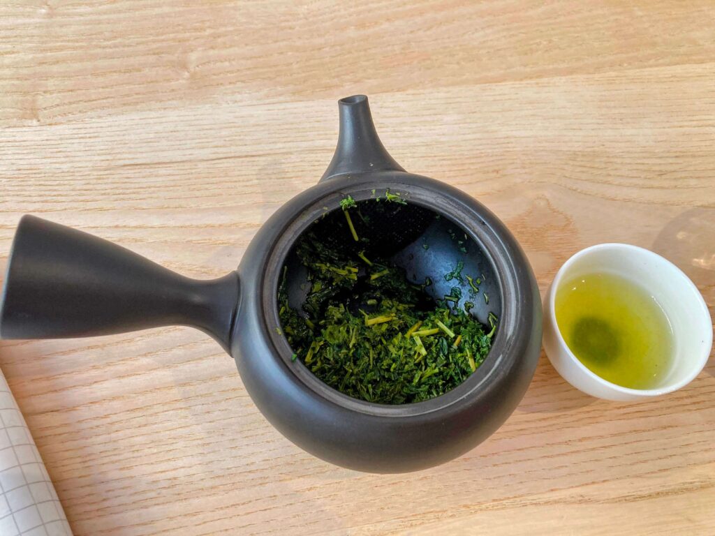 A black Japanese teapot with used Sayama Sencha leaves inside sits on a light wooden table beside a small white cup containing a small amount of green tea—perfect for Japanese tea tasting.