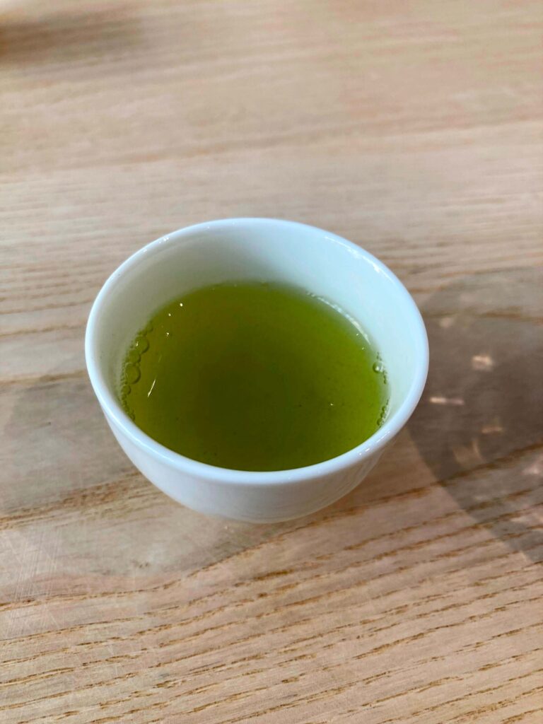 A small white cup filled with bright Sayama Sencha tea sits on a light wooden surface.