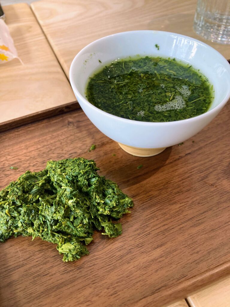 A white bowl filled with green herbal tea, likely Sayama Sencha, sits on a wooden surface next to wet, chopped green tea leaves. A glass of water and wooden tray are visible in the background, evoking a Japanese Tea Tasting experience.