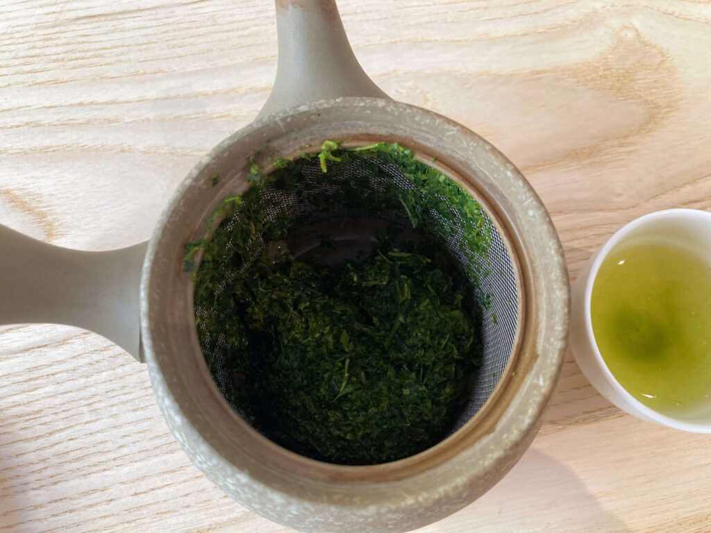 A teapot with used Sayama Sencha green tea leaves in a metal strainer sits next to a small cup filled with light green tea, all placed on a light wooden surface. Perfect for Japanese tea tasting moments.